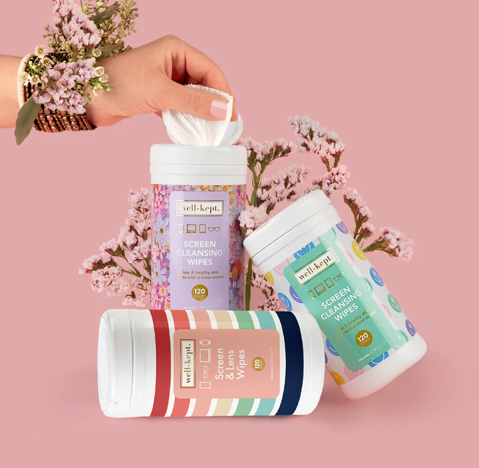 Three Well-Kept screen cleaning wipes canisters in different patterns, with a hand pulling out a wipe, styled with flowers on a pink background