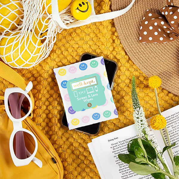 All Smiles print Well-Kept screen and lens wipes pack styled with sunglasses, a straw hat, a yellow tote bag, an open book, and flowers