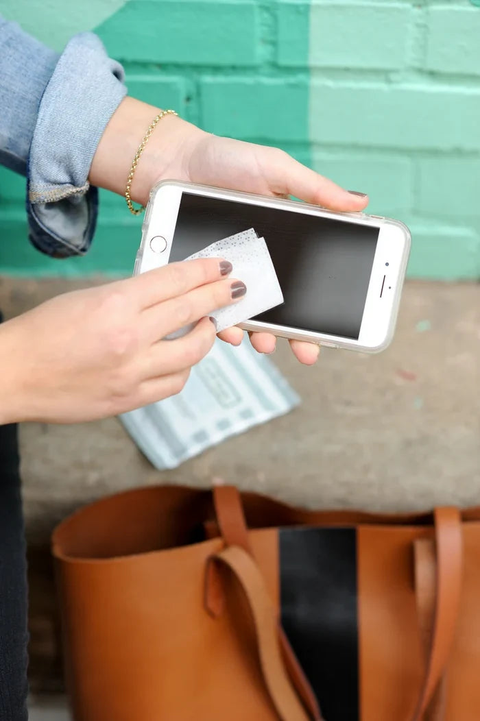Hand cleaning a phone screen with a Well-Kept wipe, next to a leather tote bag against a teal and white brick wall