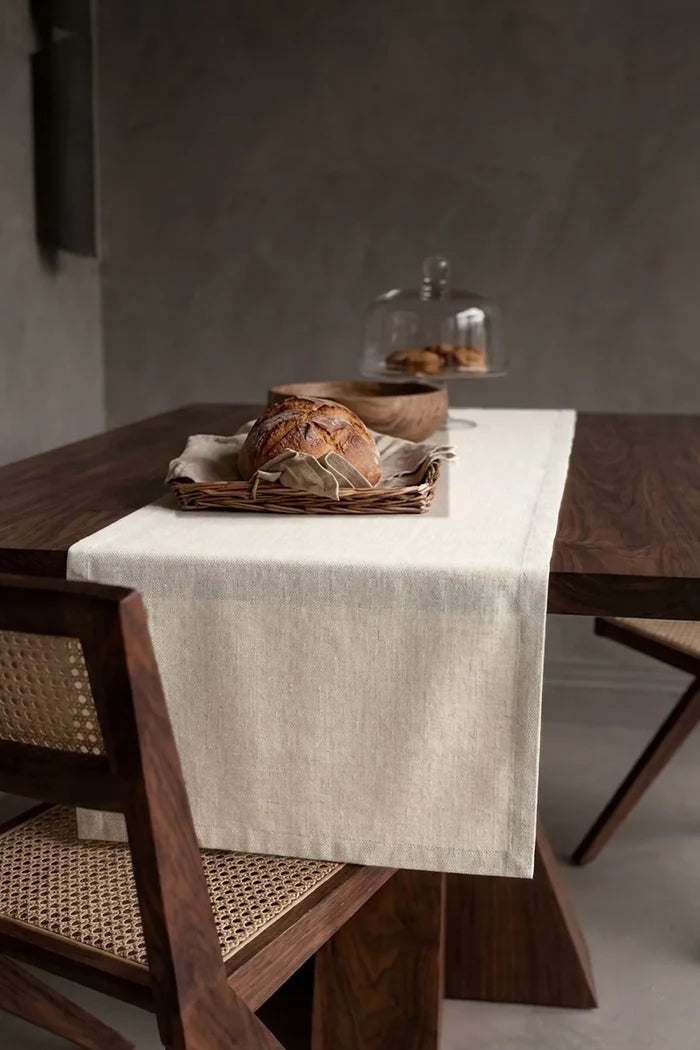 Natural plain linen cotton blend table runner draped over a walnut dining table with a bread basket and wooden bowls. 16 x 51 in, handmade in Denizli, OEKO-TEX. VivaMaison.