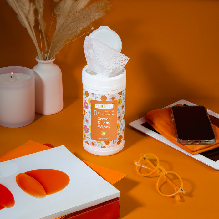 Floral print Well-Kept screen and lens wipes canister with a wipe pulled out, styled on orange with a candle, sunglasses, a phone, and a makeup sponge