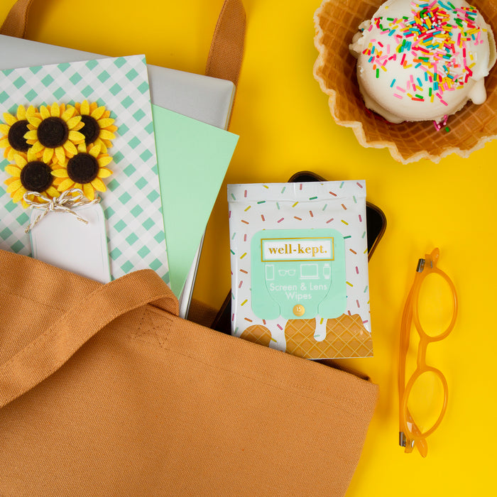 Ice cream sundae and sprinkles print Well-Kept screen and lens wipes pack in a mustard tote bag with sunflowers, notebooks, and glasses on a yellow background